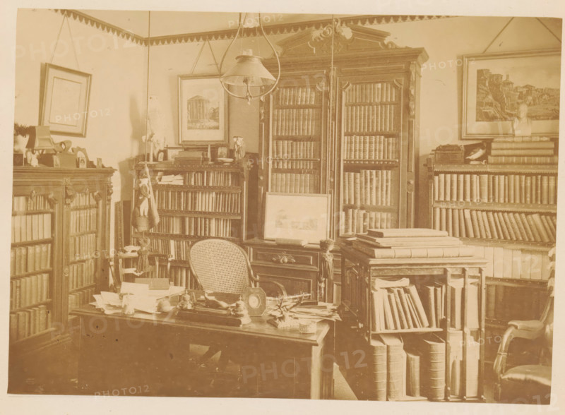 Memorial of William R. Dimmock, master of Adam Academy, photograph, page, Quincy, c. 1873 - in or before 1878, photographer, North America, Massachusetts, paper, albumen print, height 126 mm x width 178 mm, learned society, academy of sciences, anonymous historical persons depicted in a group, in a group-portrait, façade (of house or building), A sepia-toned room with tall wooden bookshelves filled with books, some with glass-fronted cabinets and ornate carved tops; a large central cabinet with double doors and shelves of books stands against the back wall, flanked by framed pictures and a bust on a shelf; a desk in the foreground cluttered with papers, small objects, and a tabletop clock, with a cane-backed chair pushed close to it; stacks of large books and rolled materials sit on a smaller table and open shelving beside the desk; a pendant light hangs from the ceiling above the central area and decorative trim runs along the top of the walls; various framed artworks hang on the wall...