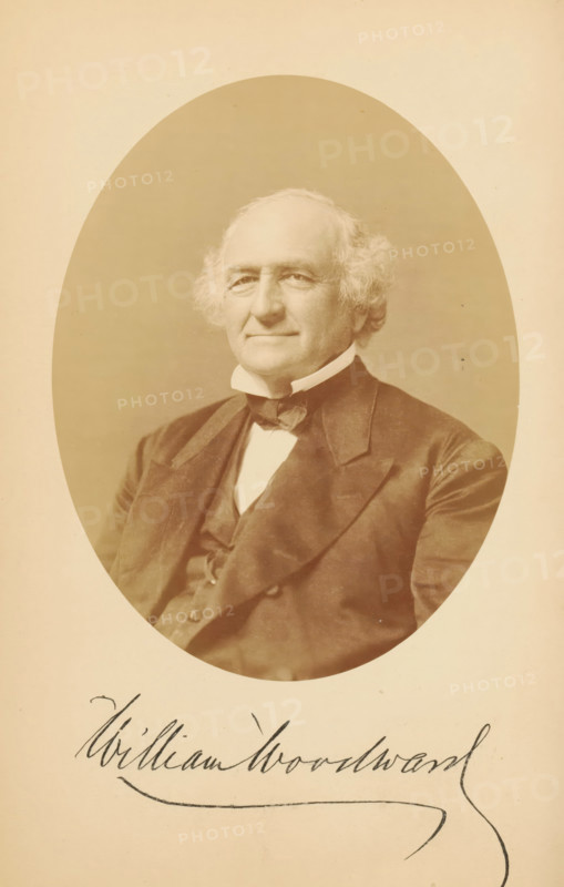 Portrait of William Woodward, William Woodward, photograph, page, photographer: Bendann Bros., c. 1866 - in or before 1871, paper, albumen print, height 147 mm x width 114 mm, Person, historical persons, An oval sepia-toned portrait of an older man with light wavy hair and sideburns, wearing a dark suit jacket, a vest, a high white collar, and a bow tie, centered on a light background with handwritten cursive text partially visible below the portrait.