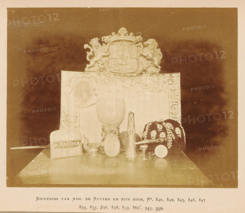 Collection of souvenirs of Admiral Michiel de Ruyter and his son, Souvenirs van Adm. de Ruyter en zijn zoon, photograph, page, number: 'No. 840, 842, 843, 846, 847, 853, 855, 856, 859, 860*, 945, 952.', photographer: Pieter Oosterhuis, Amsterdam, 1876, paper, albumen print, albumendruk, height 127 mm x width 162 mm, glass, rummer, container of precious metal, medal, badge of a guild, Historische Tentoonstelling Amsterdam, Oudemannenhuis (Amsterdam), Period, A sepia-toned photograph shows a tabletop display of objects arranged in front of an ornate backdrop topped by a decorative crest flanked by a lion and a horse. In front of the backdrop a large goblet-like vessel sits centrally, accompanied by a tall narrow spout vessel and a smaller rounded vessel nearby. To the right of the vessels sits a dark fabric cap or cushion decorated with light floral embroidery and trim, with a cylindrical rod resting beside it. To the left a rectangular block with an inscribed band is visible next to a s...