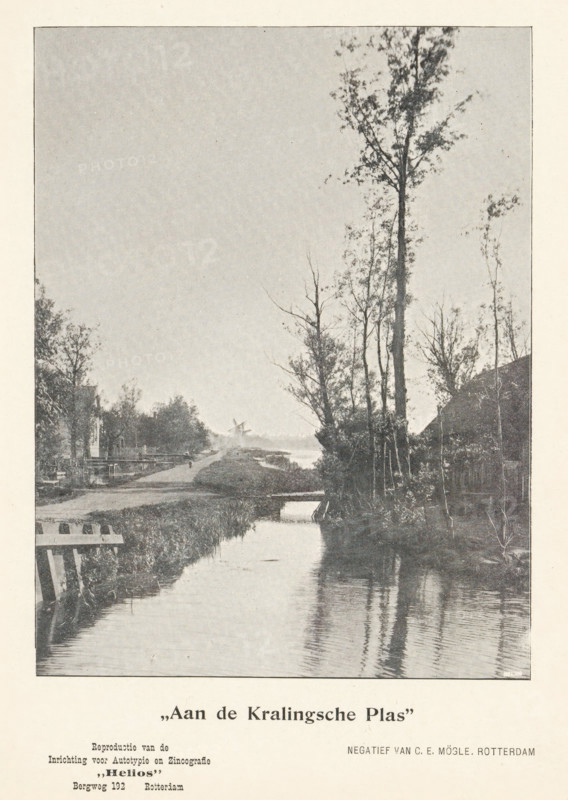 View of the Kralingse Plas, 'At the Kralingse Plas', page, photomechanical print, inscription: 'Reproduction of the, Establishment for Autotyping and Zincography, 'Helios', Bergweg 192 Rotterdam', photographer: Carl Emil Mögle, Rotterdam, cliché maker: Helios (Rotterdam) (possibly), c. 1895 - in or before 1900, printer (person), paper, autotypie, height 151 mm x width 114 mm, brook, Kralingse Plas, A vertical black-and-white photograph showing a narrow waterway running from the foreground toward the background, with reflections of tall, thin trees on the water surface. On the right bank a group of tall trees rises above a darker area of vegetation and a sloped structure partly visible behind them. The left bank shows low grassy edges, a wooden railing or fence near the foreground, and additional trees and small structures receding into the distance. A distant windmill silhouette is visible on the horizon where the waterway narrows, and the sky above is largely clear. The image is borde...