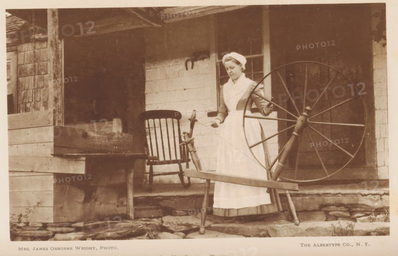 Woman spins yarn, In ye olden time, page, photomechanical print, photographer: James Osborne Wright, cliché maker: The Albertype Company, 1887 - 1907, paper, collotype, photoprint, height 109 mm x width 177 mm, Print on second unnumbered page after page 268., historical persons - BB - woman, spinning (manufacture of yarn), A sepia-toned photograph of a woman standing on a wooden porch holding a large spinning wheel, wearing a long dress with a light apron and a head covering, with one hand on the wheel and the other holding fibers; beside her is a wooden chair with a slatted back and a small spinning apparatus mounted nearby; the porch shows weathered wooden siding, posts, and a stone step, and a wooden box or trough sits to one side.