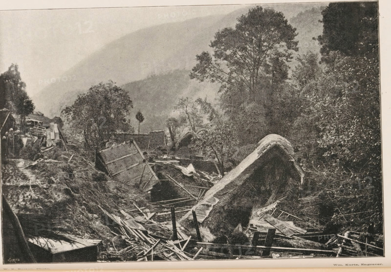 View of the devastation in a village after an earthquake in Japan, Effects of the great earthquake in Japan, page, photomechanical print, inscription: 'Kurtz', photographer: William Kinnimond Burton, Japan, cliché maker: Kurtz, 1887 - 1907, paper, autotypie, height 115 mm x width 172 mm, consequences of an earthquake, A black-and-white photograph showing a landscape with broken wooden structures and scattered debris in the foreground, including planks, beams, and tilted panels partially buried in earth and vegetation. A large curved mound covered in thatch or similar material rises amid the wreckage. Trees with dense foliage stand behind the ruined area, and taller trees extend upward toward a hazy, sloping hillside in the background. On the left edge, a few small figures stand near more damaged wooden constructions and foliage. The scene is framed by a narrow border at the top and bottom.