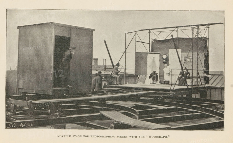 Movable Stage for Photographing Scenes with the 'Mutograph', Page, photomechanical print, inscription: 'SCI. AM. NY', maker: unknown, c. 1892 - in or before 1897, paper, autotypie, height 104 mm x width 191 mm, the stage and its equipment, A large outdoor scene on a raised platform shows a corrugated rectangular structure with an open doorway on the left and a scaffolding-like frame on the right supporting a curtained backdrop and a small enclosed area. Several people are present: one person leaning out from the doorway of the corrugated structure, a few individuals standing or seated on the platform near the frame, and two seated figures inside the framed area near the backdrop. The platform rests on visible metal beams and rails with wheels and mechanical elements beneath and around it. A plank crosses part of the platform in the foreground, and a tall angled pole or boom rises from near the center toward the right. The overall image has a grainy, monochrome appearance.