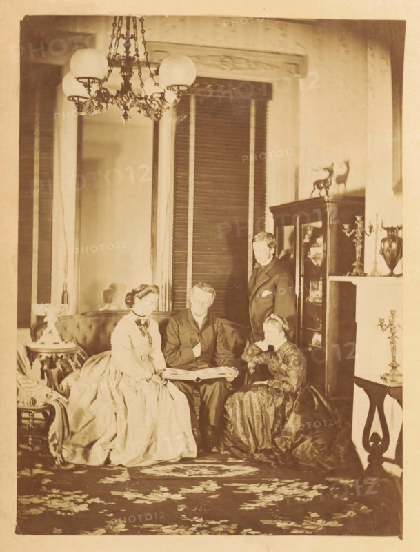 Two men and two women look at a photo album in a living room, photograph, page, photographer: JC Browne, c. 1861 - in or before 1866, paper, albumen print, albumendruk, height 167 mm x width 125 mm, achter semi-transparent cover., adult woman, adult man, living-room, parlour, sitting-room, A sepia-toned interior scene showing four people arranged around a low table, with two women seated on a curved upholstered sofa, a man seated between them holding open a large book or album, and another man standing behind them near a glass-fronted cabinet; the seated women wear full, floor-length dresses with fitted bodices and the men wear dark suits; the room includes a patterned rug, ornate chandelier with globe shades hanging from the ceiling, decorative wood trim and tall shutters behind the group, a display cabinet topped with small stag figurines, a mantel with candelabra and a vase, a small round side table with a decorative lamp, and additional furniture and ornamentation visible around th...