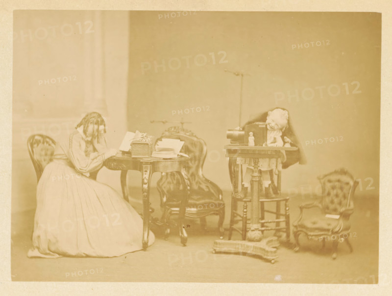 A little boy plays at a table, while a girl reads at another table, photograph, page, photographer: James Inglis, c. 1861 - in or before 1866, paper, albumen print, height 115 mm x width 157 mm, behind semi-transparent cover., girl (child between toddler and youth), toddler (male), A sepia-toned indoor scene showing a woman seated and leaning her head on her hand next to an ornate round wooden table with papers and a decorative box on it, upholstered chairs around the table, a small wooden stand holding a camera and a doll positioned behind the camera, another small upholstered chair to the right with a doll sitting on it, and various small objects and fabric on the floor in front of the stand, all set against a plain wall.