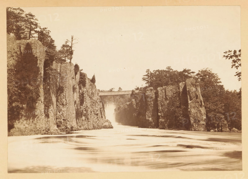View of the Great Falls of the Passaic River, New Jersey, photograph, page, photographer: J. Reid, Passaic, c. 1861 - in or before 1866, North America, paper, albumen print, height 117 mm x width 166 mm, behind semi-transparent cover., waterfall, A sepia-toned landscape showing a river flowing between tall vertical rock cliffs, with water appearing smooth and blurred in the foreground, rocky faces rising steeply on both sides, trees and vegetation atop and along the cliffs, and a narrow bridge spanning the gap near the upper center of the scene.