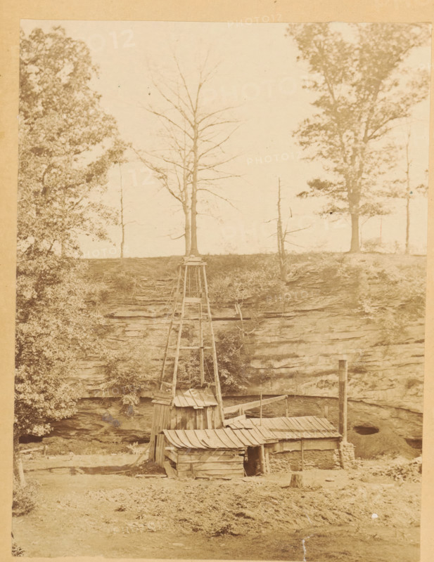 View of an oil well, photograph, page, photographer: Rogers, SG & TW, North and Central America (possibly), c. 1861 - in or before 1866, paper, albumen print, height 156 mm x width 119 mm, behind semi-transparent cover, pumping of oil, A sepia-toned photograph showing a weathered wooden structure with a slanted corrugated roof and rough plank walls situated at the base of a rocky cliff, a tall wooden framework or derrick rising from the structure with visible crossbeams and supports, a narrow deck or platform extending alongside the building, a vertical metal pipe or smokestack near the right side of the structure, layered rock faces forming the cliff behind with small ledges and vegetation, trees lining the top of the cliff including a mix of leafed and bare-branched trunks, dense foliage visible along the left edge, and a grassy foreground with a small stump and scattered ground cover.