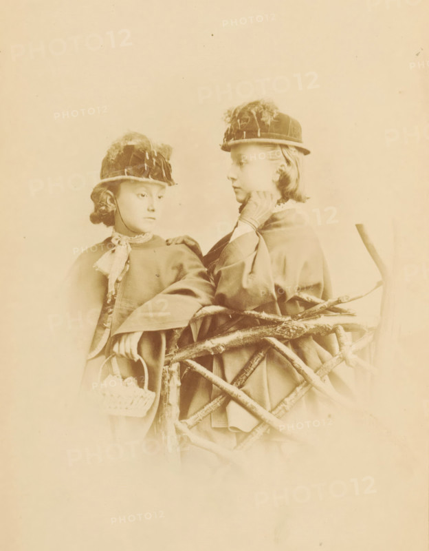 Portrait of two unknown young girls with hats, photograph, page, photographer: William Notman, c. 1861 - in or before 1866, paper, albumen print, height 157 mm x width 115 mm, behind semi-transparent cover., historical persons not known by name - BB - woman, girl (child between toddler and youth), Two young girls wearing round hats with decorative feathers stand behind a rustic wooden fence made of intertwined branches, both dressed in cloaks with high collars and one girl holding a small woven basket; one girl looks slightly downward while the other faces her with a hand near her neck, and the scene is presented in a soft sepia tone.
