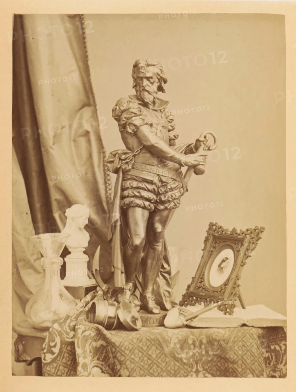 Bronze sculpture, photo frame, pipe and other items on a table, photograph, page, photographer: Frederick A. Wenderoth, c. 1861 - in or before 1866, paper, albumen print, height 166 mm x width 125 mm, behind semi-transparent cover., sculpture, pipe, tobacco, A photograph of a tabletop still life dominated by a tall statue of a bearded man in ornate period clothing holding a ring and sword, standing on a pedestal, with a draped patterned cloth beneath; to the left of the statue there is a glass vessel with a wide base and a smaller bust on a plinth, and a tall folded fabric backdrop; to the right of the statue an ornate framed oval portrait rests on a small easel, and a smoking pipe lies on the table nearby; scattered around the base of the statue are small metal cups and a rolled textile, and decorative elements and fabric textures fill the foreground.