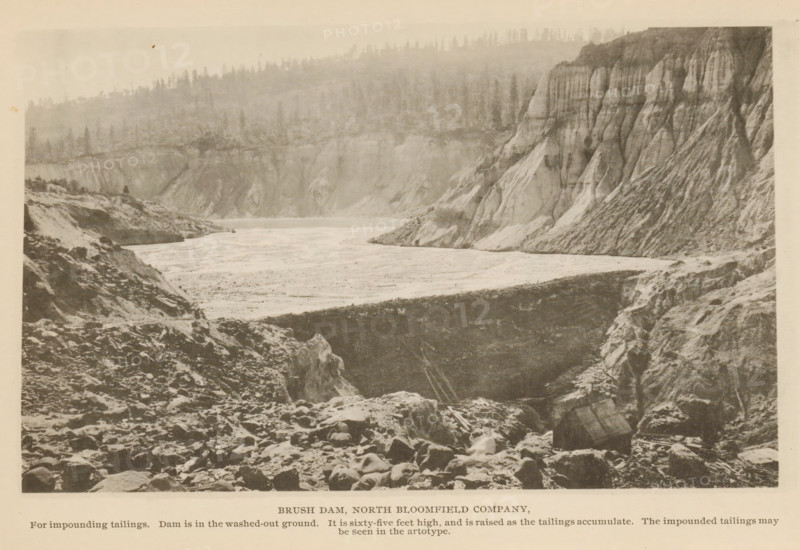 View of Brush Dam in North Bloomfield, California, Brush Dam, North Bloomfield Company, page, photomechanical print, inscription: 'For impounding tailings. Dam is in the washed-out ground. It is sixty-five feet high, and is raised as the tailings accumulate. The impounded tailings may, be seen in the artotype.', North Bloomfield, c. 1885 - in or before 1890, North America, paper, collotype, photoprint, height 110 mm x width 180 mm, mine, A sepia-toned landscape photograph showing a wide river or reservoir stretching between steep, rocky banks with layered cliffs on one side and a rugged, rubble-strewn foreground on the other, the shoreline appearing uneven and eroded, with a high rim of cliffs displaying vertical striations and sloping faces, a treeline visible along the far ridge above the cliffs, scattered rocks and boulders in the foreground, and a generally overcast or hazy sky.