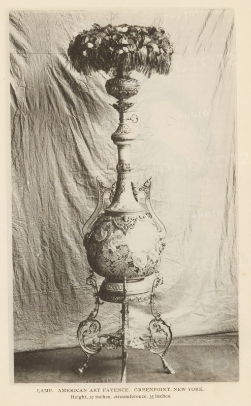 Lamp. American Art Fayence. Greenpoint, New York, Decorated lamp, page, photomechanical print, inscription: 'Height, 57 inches, circumference, 35 inches.', c. 1885 - in or before 1890, North America, paper, collotype, photoprint, height 183 mm x width 110 mm, lighting, lamps, An ornate floor lamp with a decorative base and stand is photographed against a wrinkled fabric backdrop, showing a detailed, rounded central body with floral reliefs and curved handles, a tall neck with carved sections and ornamental knobs, a wide fringed shade topped with clustered floral elements, and an elaborate tripod-like metal support with scrollwork feet.