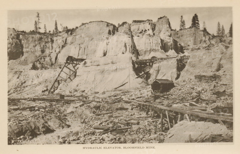 Hydraulic Elevator, Bloomfield Mine, View of a hydraulic elevator in the Bloomfield Mine, California, page, photomechanical print, North Bloomfield, c. 1885 - in or before 1890, North America, paper, collotype, photoprint, height 110 mm x width 183 mm, mine, A sepia-toned photograph of a rugged landscape with steep, light-colored cliff faces rising across the background, punctuated by scattered tall coniferous trees along the ridge. In front of the cliffs are remnants of wooden industrial structures, including angled supports and a long elevated timber chute extending up the slope and across a gap, with a small dark wooden building situated near the base of the cliffs. The foreground shows rough, rocky terrain strewn with debris and timber, and a raised wooden track or walkway running horizontally toward the right side of the image. The scene has a weathered, aged appearance.