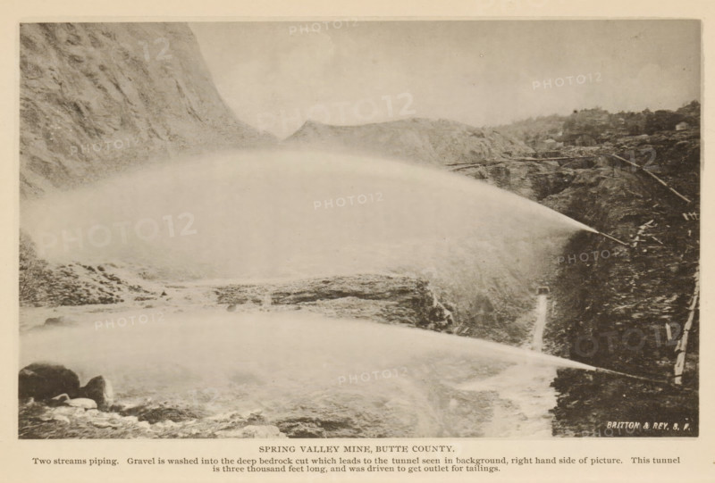 View of Spring Valley Mine in Butte County, California, Spring Valley Mine, Butte County, page, photomechanical print, inscription: 'Two streams piping. Gravel is washed into the deep bedrock cut which leads to the tunnel seen in background, right hand side of picture. This tunnel, is three thousand feet long, and was driven to get outlet for tailings.', maker: Britton & Rey, c. 1885 - in or before 1890, North America, paper, collotype, photoprint, height 112 mm x width 183 mm, mine, A sepia-toned landscape photograph showing steep rocky cliffs on both sides of a wide, misty plume of water or spray arcing across the scene from the right toward the left, with rough rocky terrain and scattered boulders visible beneath and around the plume, a narrow ledge or path along the right cliff with some wooden structures or rails, distant low hills visible through the mist in the background, and a light border surrounding the image.