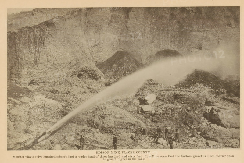 View of Hobson Mine in Placer County, California, Hobson Mine, Placer County, page, photomechanical print, inscription: 'Monitor playing five hundred miner's inches under head of three hundred and sixty feet. It will be seen that the bottom gravel is much coarser than, the gravel higher in the bank.', c. 1885 - in or before 1890, North America, paper, collotype, photoprint, height 111 mm x width 183 mm, mine, A sepia-toned photograph shows a rocky hillside with steep exposed soil and vertical streaks on the cliff face, loose rubble and piles of gravel scattered across the slope, a large arcing spray of water originating from a pipe or nozzle near the lower left and extending toward the upper right where mist obscures part of the terrain, several men standing and working among the rocks near the bottom edge, and scattered larger rocks and boulders among finer debris.