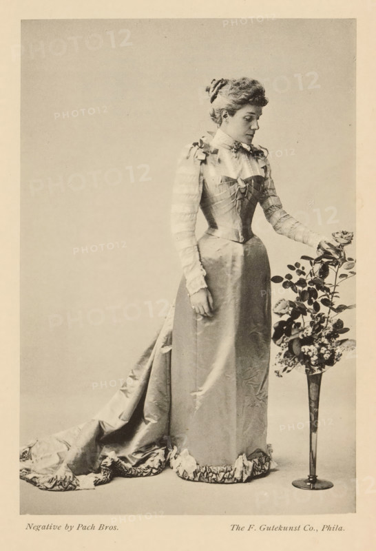 The infanta Eulalia, page, photomechanical print, photographer: GW Pach, cliché maker: Gutekunst & Co., F., Philadelphia, c. 1888 - in or before 1893, North America, paper, collotype, photoprint, height 155 mm x width 106 mm, Person, Eulalia van Bourbon, ruler, sovereign - BB - female ruler, A sepia-toned photograph shows a woman standing in profile with her head slightly turned downward toward a tall slender vase of flowers beside her. She is wearing a long, satin dress with a fitted bodice, puffed sleeves with horizontal bands, and a high collar adorned with a small decoration; the skirt extends into a ruffled train on the floor. Her hair is styled up and held with a ribbon or fabric accessory. One of her hands rests at her side while the other reaches toward a rose in the vase, which contains a stem with leaves and additional blooms and foliage. The background is plain and light, and the image has a narrow border around it.