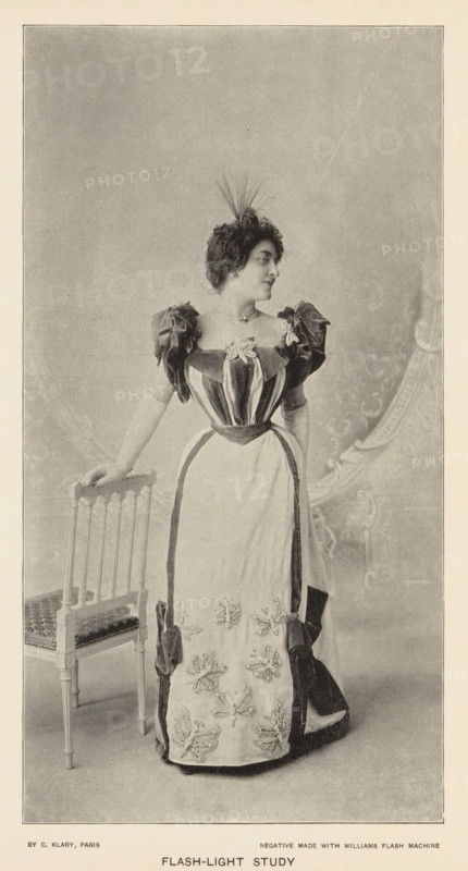 Flash-light study, page, photomechanical print, photographer: Charles Louis Klary, Paris, cliché maker: anonymous, c. 1890 - in or before 1895, paper, autotypie, height 192 mm x width 101 mm, adult woman, studio, A sepia-toned portrait of a woman standing next to a wooden chair, wearing a floor-length dress with a fitted, striped bodice and puffed sleeves, a contrasting apron-like front panel adorned with floral embellishments, and large bows at the hips; she has short curly hair topped with a small feathered headpiece and wears a necklace, her left hand resting on the back of the chair while she looks to her right, and a patterned backdrop is visible behind her.