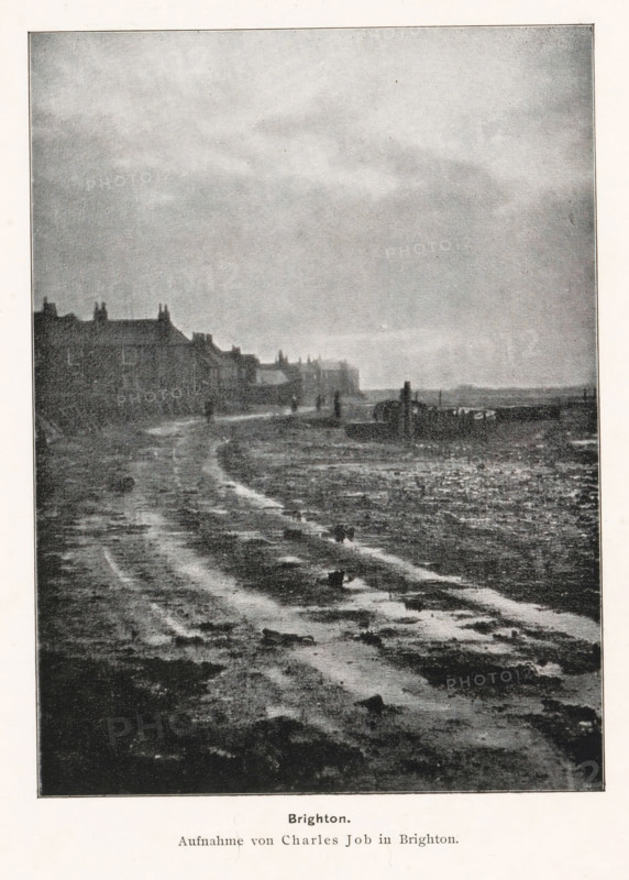 Brighton, View of the Brighton coast, page, photomechanical print, photographer: Charles Job, cliché maker: anonymous, printer (person): Wilhelm Knapp Verlag, Halle an der Saale, c. 1893 - in or before 1898, printer (person), Saxony-Anhalt, paper, autotypie, height 162 mm x width 120 mm, coast, A grainy black and white scene shows a wide muddy shoreline with visible wet ruts and patches of darker mud leading toward the horizon, a row of low buildings with pitched roofs and chimneys lines the left side, several small human figures stand or walk near the distant buildings, a faint line of structures and a low pier or breakwater extend into the water on the right, and a broad overcast sky fills the upper portion of the image.