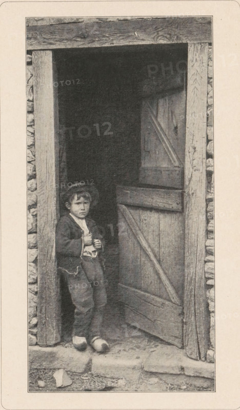 Boy in clogs in a doorway, page, photomechanical print, photographer: Paul Bourgeois, Paris, cliché maker: anonymous, printer (person): Wilhelm Knapp Verlag, c. 1893 - in or before 1898, printer (person), paper, autotypie, height 178 mm x width 89 mm, boy (child between toddler and youth), open door, wooden shoes, clogs, A sepia-toned photograph shows a young child standing in a wooden doorway of a rustic structure with stone-filled walls; the child wears a dark jacket over a lighter shirt, loose trousers, and simple shoes, and a hat sits on their head while they hold an object close to their chest with both hands; the partially open wooden door beside them has visible planks and diagonal bracing, and the door frame and surrounding stones show rough texture and weathering, with a stepped stone threshold and scattered small stones and debris on the ground outside.