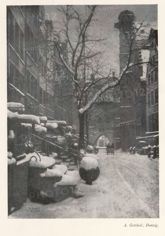 View of a street in the snow, page, photomechanical print, photographer: Albert Gottheil, cliché maker: Meisenbach, Riffarth & Cie. (possibly), Berlin, 1902 - 1903, 1902, paper, autotypie, height 179 mm x width 127 mm, Print on first unnumbered page after page XI., street, snow, A narrow snow-covered street flanked by tall buildings with rows of stoops and rounded stone posts topped with snow, a large leafless tree with snow on some branches standing near the center, a distant arched gateway at the end of the street, faint outlines of people and a horse-drawn vehicle near the gateway, and a tall chimney-like structure rising on the right side.