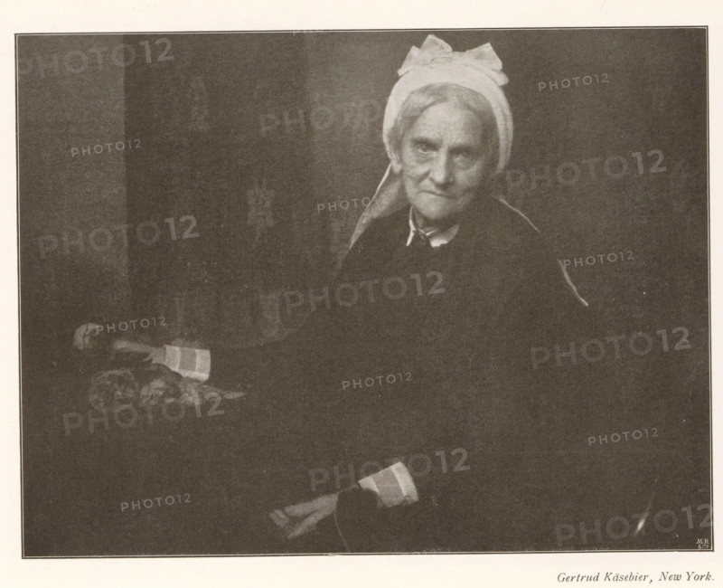 Portrait of an unknown old woman, page, photomechanical print, photographer: Gertrude Käsebier, cliché maker: Meisenbach, Riffarth & Cie. (possibly), Berlin, c. 1898 - in or before 1903, paper, autotypie, height 136 mm x width 179 mm, Print on fourth unnumbered page after page 182., historical persons not known by name - BB - woman, old woman, A monochrome photograph of an older woman seated against a dark background wearing a light headscarf tied in a bow and a dark garment with a visible collar, her face turned slightly toward the camera with a neutral expression, her hands resting in her lap with one hand holding a folded cloth, and a small table beside her holding a piece of folded fabric and some rounded objects.