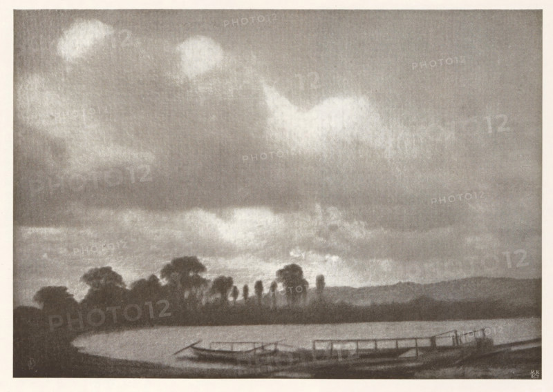 Landscape with jetty, page, photomechanical print, photographer: Otto Erhardt, cliché maker: Meisenbach, Riffarth & Cie. (possibly), Berlin, c. 1898 - in or before 1903, paper, autotypie, height 132 mm x width 190 mm, Print on second unnumbered page after page 206., landscapes, pier, quay, wharf, A grayscale landscape showing a body of water in the foreground with a curved shoreline, a small dock or platform extending over the water, and a few narrow boats moored nearby; behind the water a line of dark tree silhouettes rises against the sky, with taller slender trees interspersed among broader, rounded canopies; the sky occupies most of the image and is filled with soft, diffused clouds and lighter patches creating a textured, atmospheric backdrop.