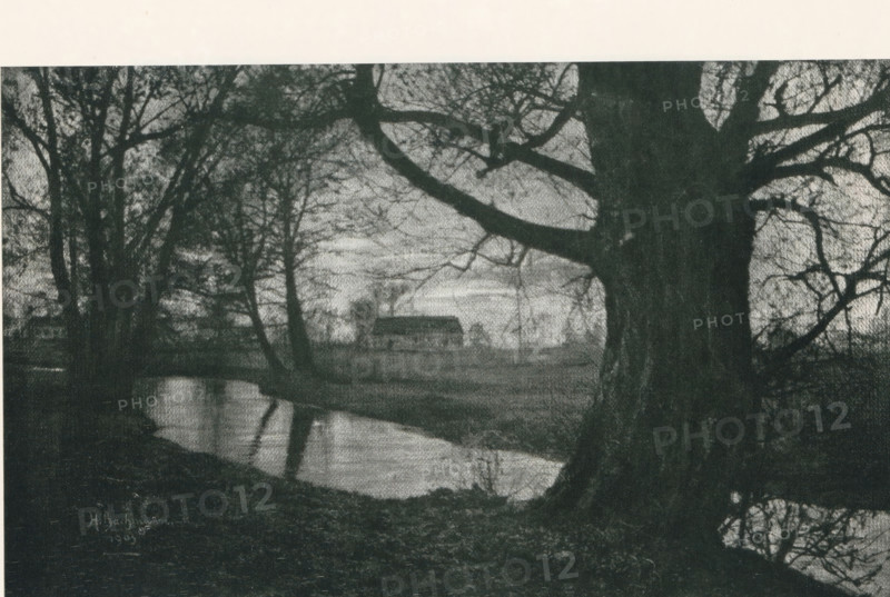 Landscape, with a house in the background, page, photomechanical print, photographer: Heinrich Bachmann, cliché maker: Meisenbach, Riffarth & Cie. (possibly), Berlin, 1903, paper, autotypie, height 134 mm x width 215 mm, Print on second unnumbered page after page 262., farm or solitary house in landscape, A wide dark-toned scene showing large tree trunks and bare branches in the foreground framing a calm reflective body of water that runs across the lower portion of the image, with the branches creating intricate silhouettes against a lighter sky; in the midground a simple rectangular building with a pitched roof sits on a flat area beyond the water, and sparse trees and low vegetation surround the building and shoreline, while the foreground ground appears uneven and textured.