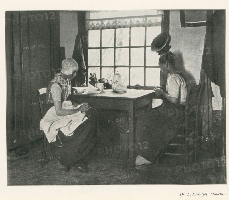 Two women sitting at a table doing needlework, page, photomechanical print, photographer: LL Kleintjes, cliché maker: anonymous, c. 1898 - in or before 1903, paper, autotypie, height 139 mm x width 174 mm, Print on second unnumbered page after page 290., adult woman, mending, repairing, clothes, A black and white scene of two seated women at a wooden table in front of a multi-paned window, one woman on the left wearing a head covering and an apron with hands in her lap, the other on the right leaning slightly forward with hands near the table edge; on the table there are a teapot, a cup, a plate, and some small items, a coat hangs on the wall beside the window, and light comes through the window panes into the room.