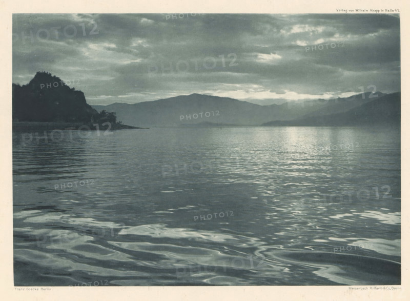 Am Luganer See, View of Lake Lugano, page, photomechanical print, photographer: Franz Goerke, Lugano, cliché maker: Meisenbach, Riffarth & Cie., Berlin, publisher: Wilhelm Knapp Verlag, Halle an der Saale, c. 1898 - in or before 1903, printer (person), Saxony-Anhalt, paper, photoaquatint, height 137 mm x width 195 mm, Print four of fourteen prints at the back the book., lake, mountains, A wide view of a calm body of water extending toward distant low mountains under a cloudy sky, with gentle ripples and reflections on the water surface, a dark silhouetted shoreline with a rocky promontory and some trees on the left, and layered mountain ridges fading into the background beneath scattered clouds.