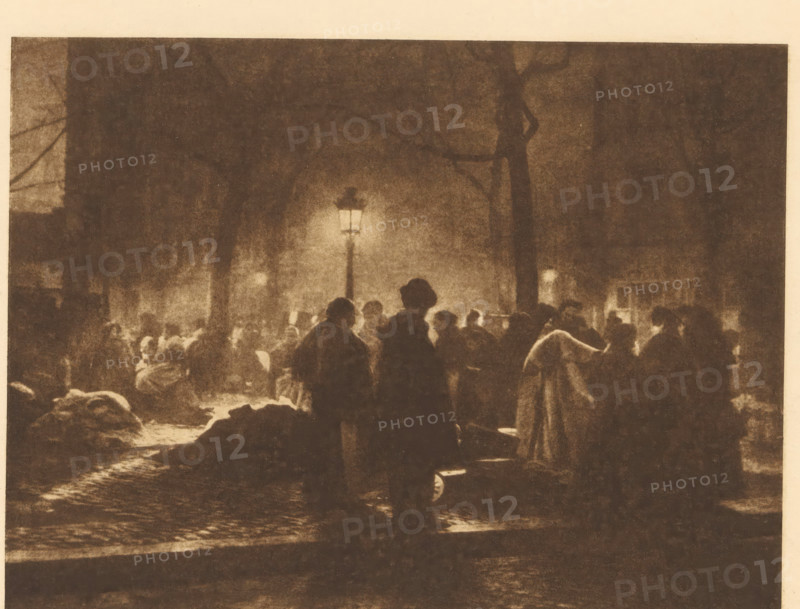View of a market in Brussels, Sonnabendmarkt in Brüssel, page, photomechanical print, photographer: A. Cumont, Brussels, cliché maker: Büxenstein & Comp., Georg, Berlin, c. 1898 - in or before 1903, printer (person), paper, photoaquatint, height 134 mm x width 180 mm, Print five of fourteen prints at the back of the book., market, A dimly lit street scene at night showing a crowd of shadowy figures gathered under a tall lit lamppost, with silhouettes of people wearing coats and hats, dark tree trunks and branches rising behind them, areas of light and shadow creating a grainy, sepia-toned atmosphere, cobblestone pavement visible beneath the figures, and blurred building shapes and windows faintly visible in the background.