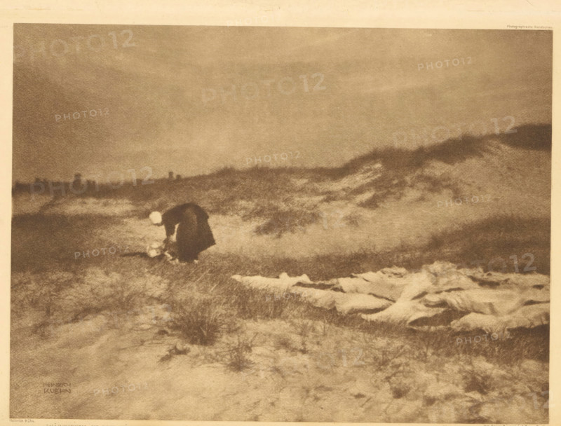 Woman washes clothes in the dunes, Wäscherin in der Düne, page, photomechanical print, photographer: Heinrich Kühn, cliché maker: Büxenstein & Comp., Georg, Berlin, c. 1898 - in or before 1903, printer (person), paper, photoaquatint, height 167 mm x width 227 mm, Print twelve of fourteen prints at the back of the book., adult woman, dunes (sea not visible), in the dunes, laundering, A grainy monochrome scene of a windswept landscape with low rolling dunes and rough grass, a solitary figure wearing a dark cloak bent forward near the ground and holding a pale round object, distant low horizon with indistinct vertical shapes, and textured foreground with scattered lighter patches on the sand.
