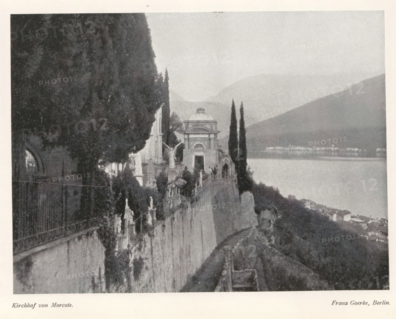 View of the Morcote cemetery, Kirchhof von Morcote, page, photomechanical print, photographer: Franz Goerke, Morcote, cliché maker: Meisenbach, Riffarth & Cie. (possibly), Berlin, c. 1898 - in or before 1903, paper, autotypie, height 149 mm x width 199 mm, churchyard, cemetery, A grayscale photograph shows a lakeside scene with a stone wall running along a steep slope, topped by a row of sculpted figures and ornate railings; tall slender trees rise near a small domed building with an arched doorway, and larger trees and foliage occupy the left side of the image; beyond the wall the calm water of a lake stretches toward distant, softly contoured hills and a small cluster of buildings along the far shore, while a narrow path follows the contour of the terrain in the foreground.