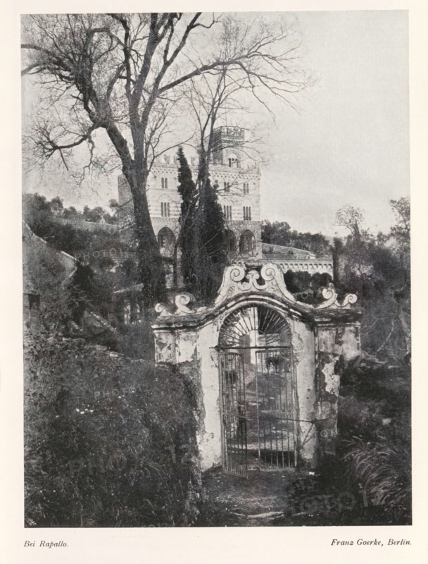 Bei Rapallo, View of a villa on a hill, near the town of Rapallo, page, photomechanical print, photographer: Franz Goerke, Rapallo, cliché maker: Meisenbach, Riffarth & Cie. (possibly), Berlin, c. 1898 - in or before 1903, paper, autotypie, height 199 mm x width 150 mm, Print on fourth unnumbered page after page 52., villa, gate, entrance, A black and white photograph showing a tall ornate stone gate with a decorative curved top and metal bars set in a weathered wall in the foreground, a narrow path leading up to the gate flanked by overgrown vegetation, a large bare-branched tree rising behind and to the left of the gate with additional leafless trees interwoven, and a multi-story building with arched windows and a crenellated tower partially visible through the trees in the midground, with distant foliage and a pale sky filling the background.