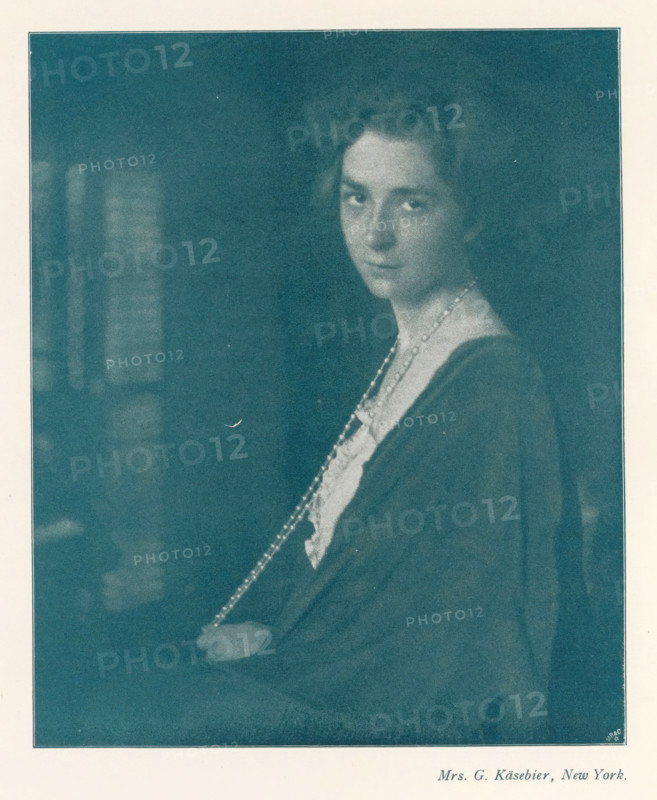 Portrait of an unknown woman with a long pearl necklace, page, photomechanical print, photographer: Gertrude Käsebier, cliché maker: Meisenbach, Riffarth & Cie. (possibly), Berlin, c. 1898 - in or before 1903, paper, autotypie, height 157 mm x width 130 mm, Print on second unnumbered page after page 80., historical persons not known by name - BB - woman, pearl, pearl necklace, A faded monochrome portrait of a woman shown from the torso upward, turned slightly to one side with her face looking toward the viewer, wearing a dark draped garment over a lighter blouse and a long beaded necklace that hangs down and is held in one hand, her hair styled away from her face, set against a dark, blurry background with a pale border surrounding the image.