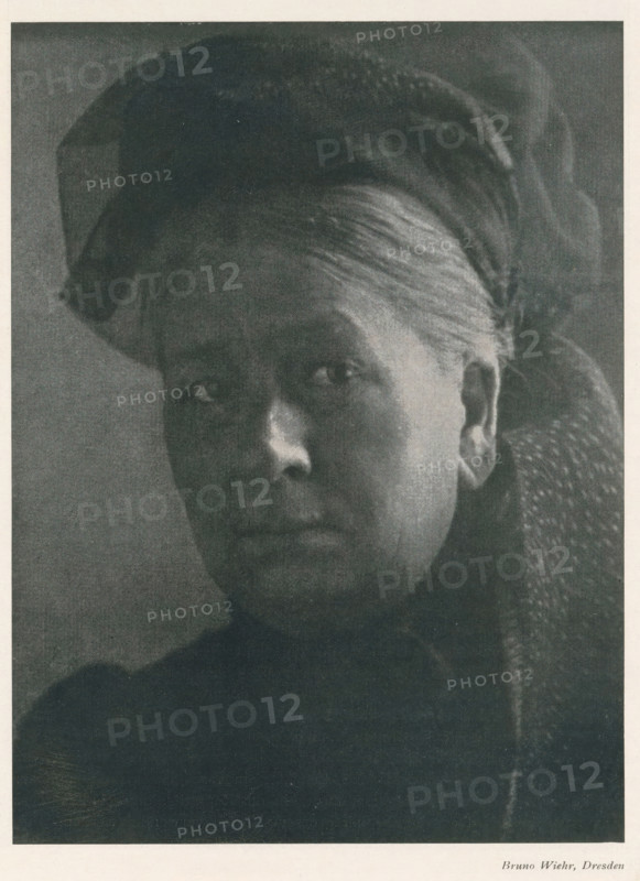 Portrait of an unknown woman, page, photomechanical print, photographer: Bruno Wiehr, cliché maker: anonymous, c. 1898 - in or before 1903, paper, autotypie, height 174 mm x width 129 mm, historical persons not known by name - BB - woman, A close-up black-and-white portrait of an older woman shown from the shoulders up, her face turned slightly toward the camera with a neutral expression, visible skin texture and fine hair pulled back under a dark head covering, one ear exposed, dark clothing with a textured fabric on one shoulder, and a plain darker background.