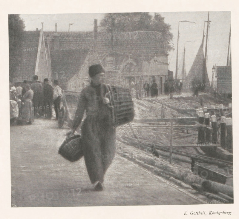 Man with two baskets in hand on a quay, page, photomechanical print, photographer: Elias Gottheil, cliché maker: Meisenbach, Riffarth & Cie. (possibly), Berlin, c. 1898 - in or before 1903, paper, autotypie, height 139 mm x width 160 mm, Print on second unnumbered page after page 132., adult man, container made of plant material other than wood: basket, A grainy black-and-white scene shows a waterfront area with a person walking toward the camera carrying a large bundle under one arm and a sack in the other hand, dressed in a long coat and a hat. In the background along the shore there are boats with tall masts and sails, stacked planks and nets, and a row of small buildings with pitched roofs. Several other people stand or walk near the buildings and along the dock, and a tree rises behind the roofs. The ground near the water appears uneven and strewn with ropes or timber, and a railing runs along the edge of the dock.