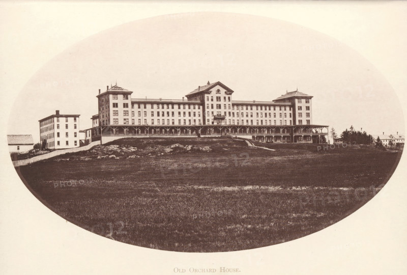 View of Old Orchard House in Old Orchard Beach, Maine, Old Orchard House, page, photomechanical print, Old Orchard Beach, c. 1875 - in or before 1880, North America, paper, collotype, photoprint, height 191 mm x width 115 mm, hotel, hostelry, inn, A wide, oval-framed sepia photograph of a large multi-story building with a long covered porch running along its front, flanked by two taller corner sections with decorative rooflines and smaller attached wings; the building sits on a slightly raised grassy, rocky slope with a fenced area and a few smaller outbuildings and houses visible to the left and right, and a strip of open lawn filling the foreground.