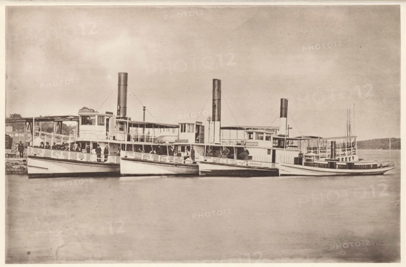 Sebenoa, Sasanoa, Samoset, Creedmoor, Eastern Steamboat Company's Bath and Boothbay Line, View of the Sebenoa, Sasanoa and Samoset and Creedmoor ferries near Bath and Boothbay, Maine, page, photomechanical print, c. 1875 - in or before 1880, North America, paper, collotype, photoprint, height 120 mm x width 184 mm, Print on second unnumbered page after page 24., ferry, A sepia-toned photograph showing a row of passenger steamships moored side by side at a dock, each vessel featuring tall smokestacks, covered upper decks, and open railings. People stand and sit along the decks and near the railings, some clustered under canopies and others near the wheelhouses. The water in front of the boats is calm and reflects parts of the hulls. In the background a low treeline and parts of the dock structure are visible. The sky above is wide and lightly textured.