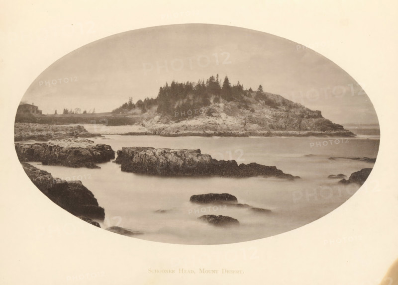 View of Schooner Head on Mount Desert Island, Maine, Schooner Head, Mount Desert, page, photomechanical print, c. 1875 - in or before 1880, North America, Mount Desert Island, paper, collotype, photoprint, height 115 mm x width 190 mm, cape (coast), A sepia-toned oval photograph depicting a rocky coastline with a foreground of dark jagged rocks partially submerged in smooth, misty water, a midground of larger rocky outcrops rising from the water, and a tree-covered promontory with dense conifer silhouettes along its ridge, a low shoreline stretching toward the left where a small building sits near the edge, and a pale sky filling the upper portion of the frame.