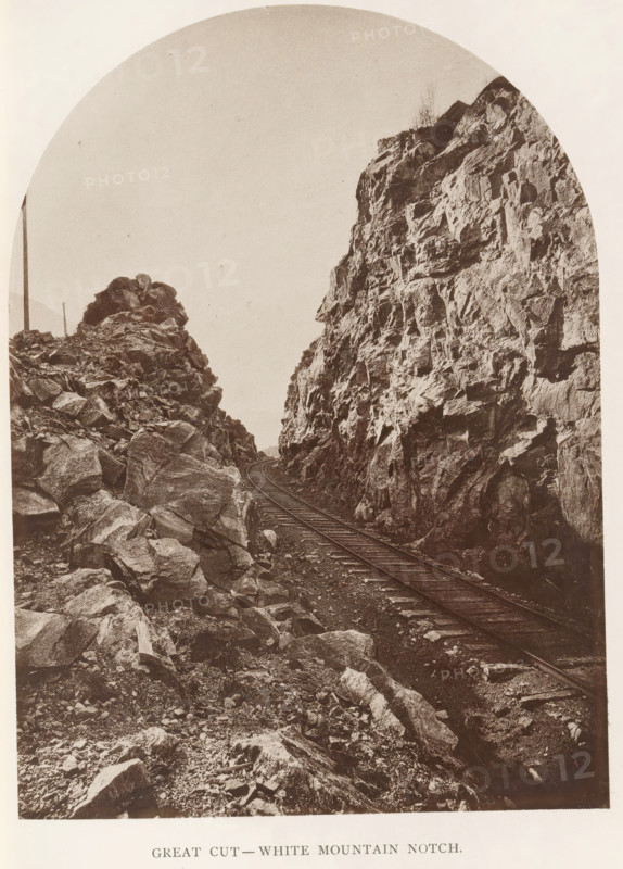 Great Cut - White Mountain Notch, View of a train track along White Mountain Notch, New Hampshire, page, photomechanical print, White Mountains, c. 1874 - in or before 1879, North America, paper, collotype, photoprint, height 173 mm, width 127 mm, Print for chapter 6., railway, train, mountains, A sepia-toned photograph showing a narrow railroad track running between two steep, rocky cliffs, with jagged rock faces and scattered boulders on the ground, the rails curving away into the distance, a tall wooden post visible near the left cliff, and sparse vegetation or small shrubs atop the cliffs under a pale sky.