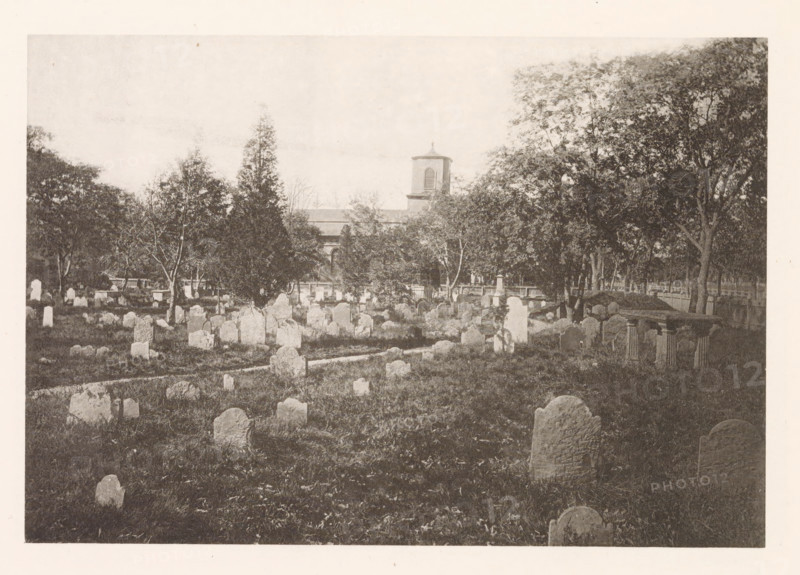 Cemetery of Christ Church in Cambridge, page, photomechanical print, photographer: William James Stillman, Cambridge (MA), cliché maker: anonymous, United States of America, c. 1866 - in or before 1876, North America, Massachusetts, paper, collotype, photoprint, height 128 mm x width 186 mm, church (exterior), churchyard (around church), A wide view of an outdoor scene showing an expanse of grass dotted with many upright stone markers of varying shapes and sizes, some rounded on top and others more irregular, arranged in rows and clusters across the foreground and middle ground, with a narrow path running among them; scattered trees of different heights stand throughout the area, their trunks and foliage creating shaded pockets, and larger trees form a dense line toward the background; beyond the trees a building with a square tower and a small roofed cupola is partially visible, its façade and roofline seen above a fence that runs along the rear of the field.