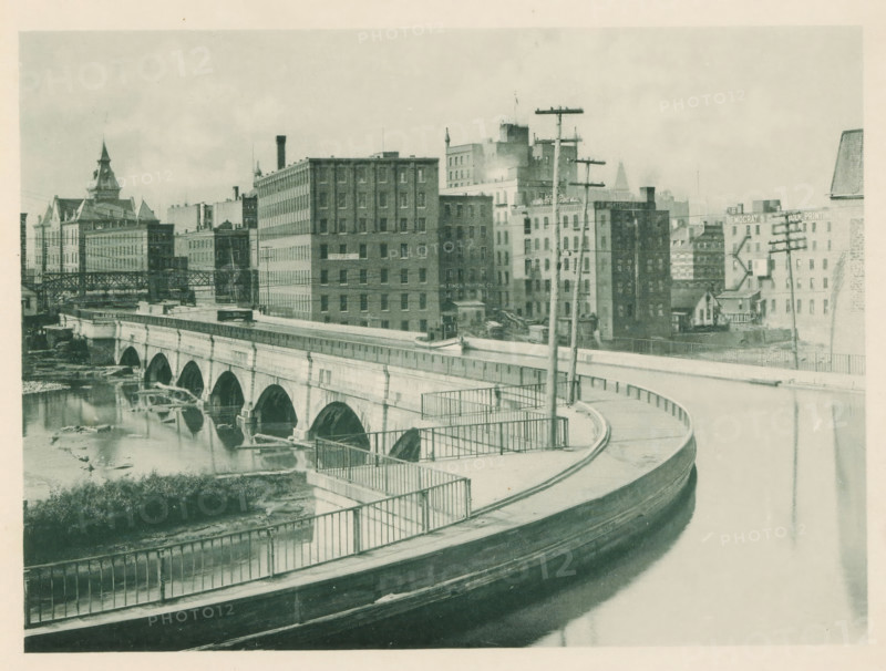 The Erie Canal viaduct at Rochester, NY, View of an aqueduct for the Erie over the Genesee in Rochester, page, photomechanical print, Rochester (New York), c. 1883 - in or before 1893, North America, paper, collotype, photoprint, height 140 mm x width 190 mm, canals, waters (in city), city-view, and landscape with man-made constructions, A curved roadway with metal railings runs along the foreground above a body of water, leading to a stone arched bridge that spans the river below; the bridge shows multiple rounded arches and reflections in the water. Beyond the bridge a cluster of multistory brick and stone buildings of varying heights lines the riverbank, with some rooftop structures, chimneys, and a tower with a pointed roof visible among them. Utility poles with crossarms and wires stand near the roadway and in front of the buildings. The riverbank shows exposed rocks and debris at the waterline, and a raised walkway or elevated structure extends across part of the scene. The photo...