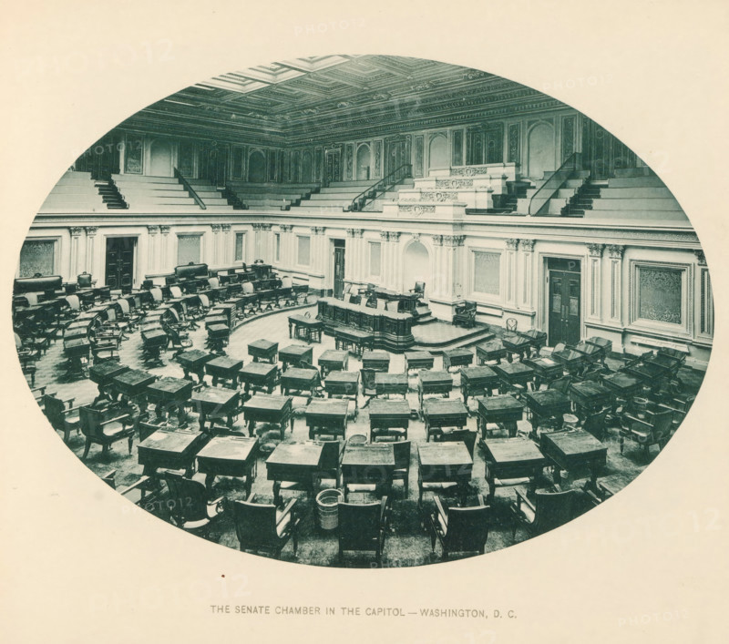Interior of the senate chamber in the Capitol in Washington DC, The senate chamber in the Capitol - Washington, DC, page, photomechanical print, Capitol (Washington DC), c. 1883 - in or before 1893, North America, Washington DC, District of Columbia, paper, collotype, photoprint, height 148 mm x width 201 mm, Upper House, Senate, national government, An oval-framed photograph of an ornate semicircular chamber with rows of wooden desks and chairs arranged in a curved formation facing a raised central dais, carved decorative paneling and fluted pilasters along the walls, an elaborately coffered ceiling, tiered gallery seating with staircases, a central lectern and benches on the dais, upholstered chairs against the walls, patterned wall panels, and various small tables and wastebaskets scattered among the desks.