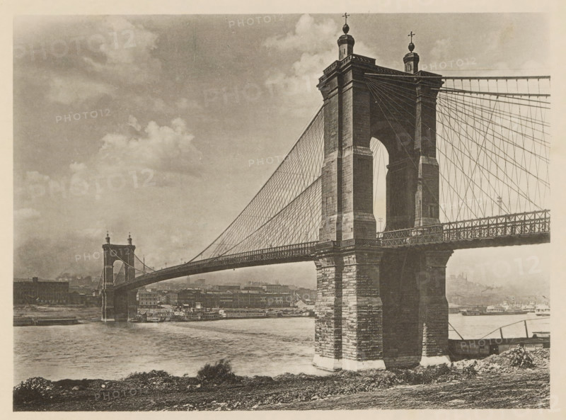 View of John A. Roebling Suspension Bridge at Cincinnati, The Ohio Suspension Bridge at Cincinnati, page, photomechanical print, Cincinnati, c. 1883 - in or before 1893, North America, paper, collotype, photoprint, height 140 mm x width 190 mm, suspension bridge, A sepia-toned photograph of a large suspension bridge spanning a river, showing a close-up of one massive stone tower with an arched opening and decorative finials on top, a network of vertical and diagonal suspension cables extending from the tower to the roadway, and the bridge deck with a latticework railing. The river flows beneath the bridge, with a vessel moored along the far bank and industrial buildings lining the waterfront beyond. The near riverbank in the foreground shows rough terrain and low vegetation, and the sky above contains scattered clouds.