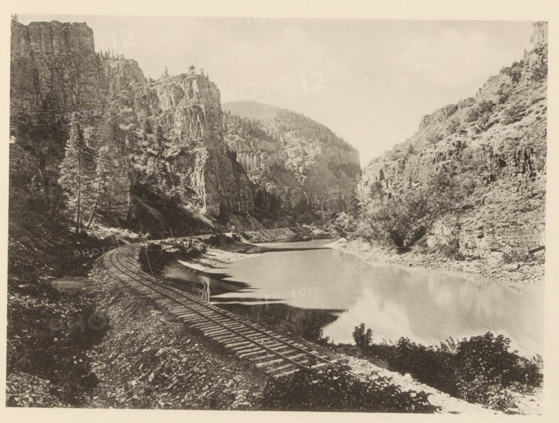 View of the Rio Grande with rock walls in Colorado, Echo Cliffs, Colorado - Denver & Rio Grande RR, page, photomechanical print, Colorado, c. 1883 - in or before 1893, North America, paper, collotype, photoprint, height 140 mm x width 191 mm, railway, train, river, inland cliff face, A sepia-toned landscape photograph showing a river running between steep rocky cliffs with trees scattered along the slopes and ridges. A curved set of railroad tracks follows the riverbank in the foreground, with wooden ties visible against a gravelly embankment. Reflections from the cliffs and vegetation appear on the calm surface of the water. The left side features a tall conifer near the base of a sheer rock face, while the right side shows terraced rock formations and scrubby vegetation. Distant hills rise behind the canyon walls beneath a pale, cloudless sky.