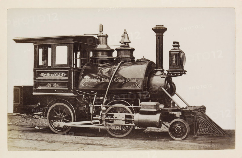 American steam locomotive, photograph, page, number: '53', United States of America, c. 1871 - in or before 1881, photographer, North America, paper, albumen print, height 124 mm x width 198 mm, Photo on page 125., steam locomotive, A sepia-toned photograph showing the side of a vintage steam locomotive with a rectangular cab featuring two windows and decorative paneling, a rounded boiler with ornate trim and raised cylindrical domes, a tall vertical smokestack, and a lantern mounted near the front. Visible mechanical linkages, pipes, and pistons run along the lower side above large spoked wheels and smaller front wheels, and a slatted metal pilot extends from the front near the rails. The locomotive rests on cobblestone pavement against a plain light background.