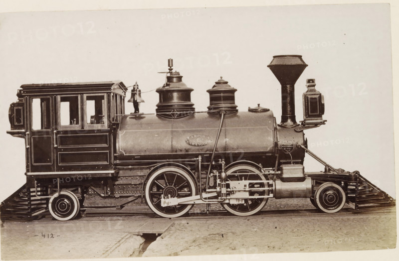 American steam locomotive, photograph, page, United States of America, c. 1871 - in or before 1881, photographer, North America, paper, albumen print, height 124 mm x width 200 mm, Photo on page 136., steam locomotive, A sepia-toned side view of an old steam locomotive showing the cab with paneled wooden sides and window openings, a rounded boiler with riveted seams and decorative trim, and large spoked driving wheels with visible connecting rods and mechanical linkages. Two domed fittings sit atop the boiler, one with a bell mounted beside it and a slender vertical whistle or valve. A tall, flared smokestack rises near the front with a rectangular lamp mounted to its side. The locomotive rests on wooden rails and a dirt surface with a small open trench visible beneath the track.