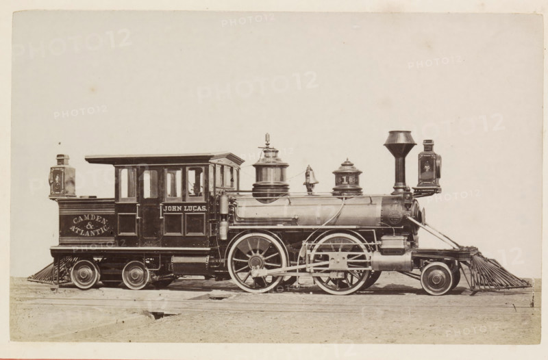 American steam locomotive, photograph, page, United States of America, c. 1871 - in or before 1881, photographer, North America, paper, albumen print, height 125 mm x width 200 mm, Photo on page 139., steam locomotive, A sepia-toned photograph of a vintage steam locomotive shown in profile on wooden tracks, featuring a rectangular cab with windows and decorative paneling, a cylindrical boiler with domed fittings and a tall funnel-shaped smokestack, large spoked driving wheels connected by visible rods and mechanical linkages, a cowcatcher at the front, lamps mounted at both ends, and various pipes, valves, and metal fittings along the body, set against an empty pale background.