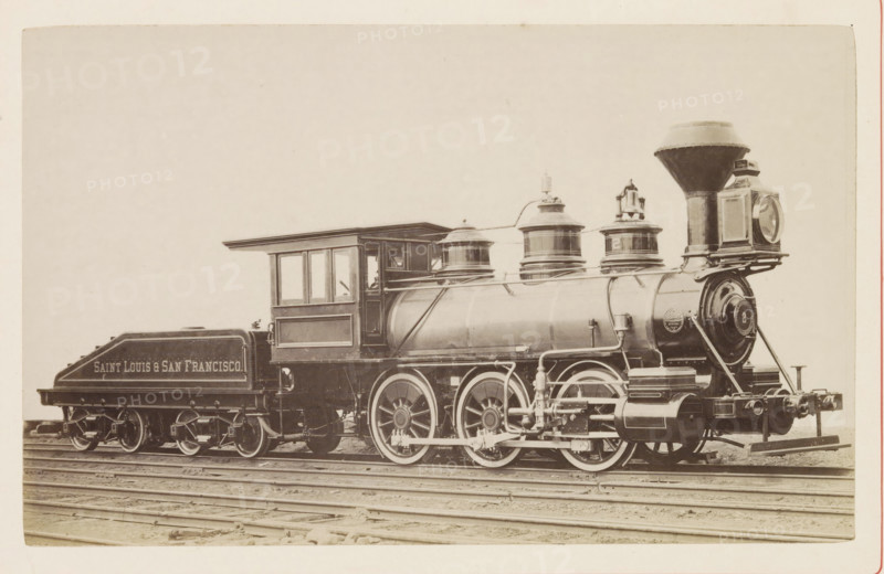American steam locomotive, photograph, page, United States of America, c. 1871 - in or before 1881, photographer, North America, paper, albumen print, height 125 mm x width 199 mm, Photo on page 146., steam locomotive, A vintage steam locomotive and its tender sit on railroad tracks against a pale background, showing a large cylindrical boiler with domed fittings and piping, a tall flared smokestack, a square headlamp mounted at the front, and a round-faced front plate. The engineer's cab has framed windows and a projecting roof, and the tender behind it has a sloped top and visible wheel assemblies. Large driving wheels with spokes and connecting rods are visible beneath the boiler, along with exposed mechanical linkages and external piping. The scene is monochrome with subtle sepia tones, and the ground beneath the tracks is visible.