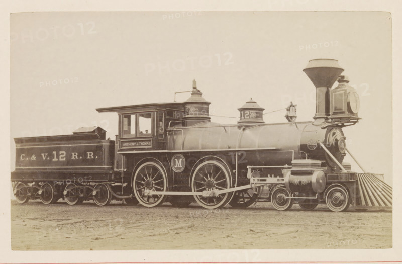 American passenger and freight steam locomotive, photograph, page, United States of America, c. 1871 - in or before 1881, photographer, North America, paper, albumen print, height 124 mm x width 199 mm, Photo on page 68., steam locomotive, A vintage steam locomotive and its attached coal car sit on flat ground, shown in a side view across the image. The locomotive has a tall funnel-shaped smokestack, a large round headlight housing at the front, a cowcatcher with angled slats, two large driving wheels with visible spokes and connecting rods, cylindrical tanks and domes along the boiler, a cab with windows and a roof overhang, and decorative round emblems on the wheels and boiler. The coal car behind the engine has a rectangular open-top shape and visible wheelsets beneath. The scene is in sepia tones and the background is plain and light, with the ground in front appearing textured and slightly uneven.