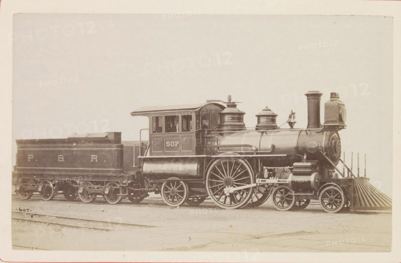 American passenger steam locomotive, photograph, page, United States of America, c. 1871 - in or before 1881, photographer, North America, paper, albumen print, height 124 mm x width 200 mm, Photo on page 78., steam locomotive, A sepia-toned image of a steam locomotive and its tender positioned on rails, showing the engine with a tall cylindrical smokestack, a rounded boiler with external piping, domed steam domes, and a cab with windows; large spoked driving wheels and smaller leading wheels are visible along with connecting rods and mechanical linkages; the front of the locomotive features a slatted pilot and a circular front plate; the attached tender has a rectangular open-top coal space and rests on a set of wheels; the scene is framed by a light background and a ground surface with visible rail tracks.